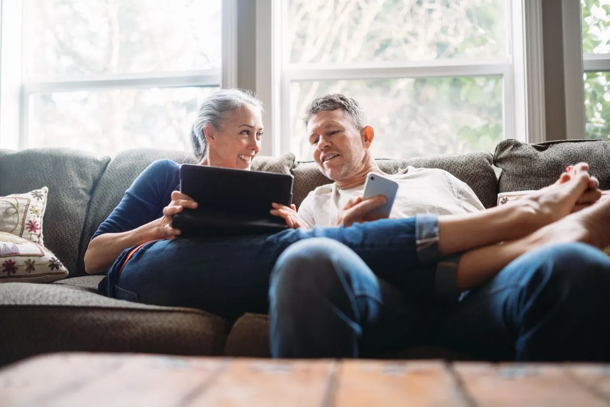 Two people sitting together on a couch, looking at a tablet and a smartphone in a bright, relaxed home setting.