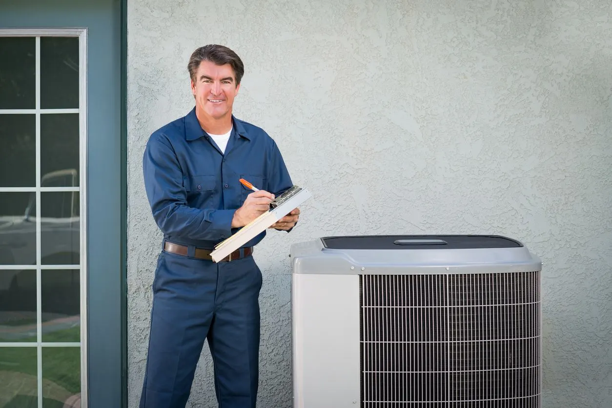 A technician in a blue uniform writes on a clipboard next to an outdoor HVAC unit against a light gray wall.