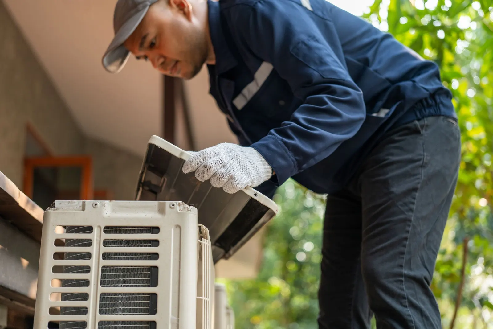 A technician in a blue uniform and gloves removes a cover from an outdoor air conditioning unit.