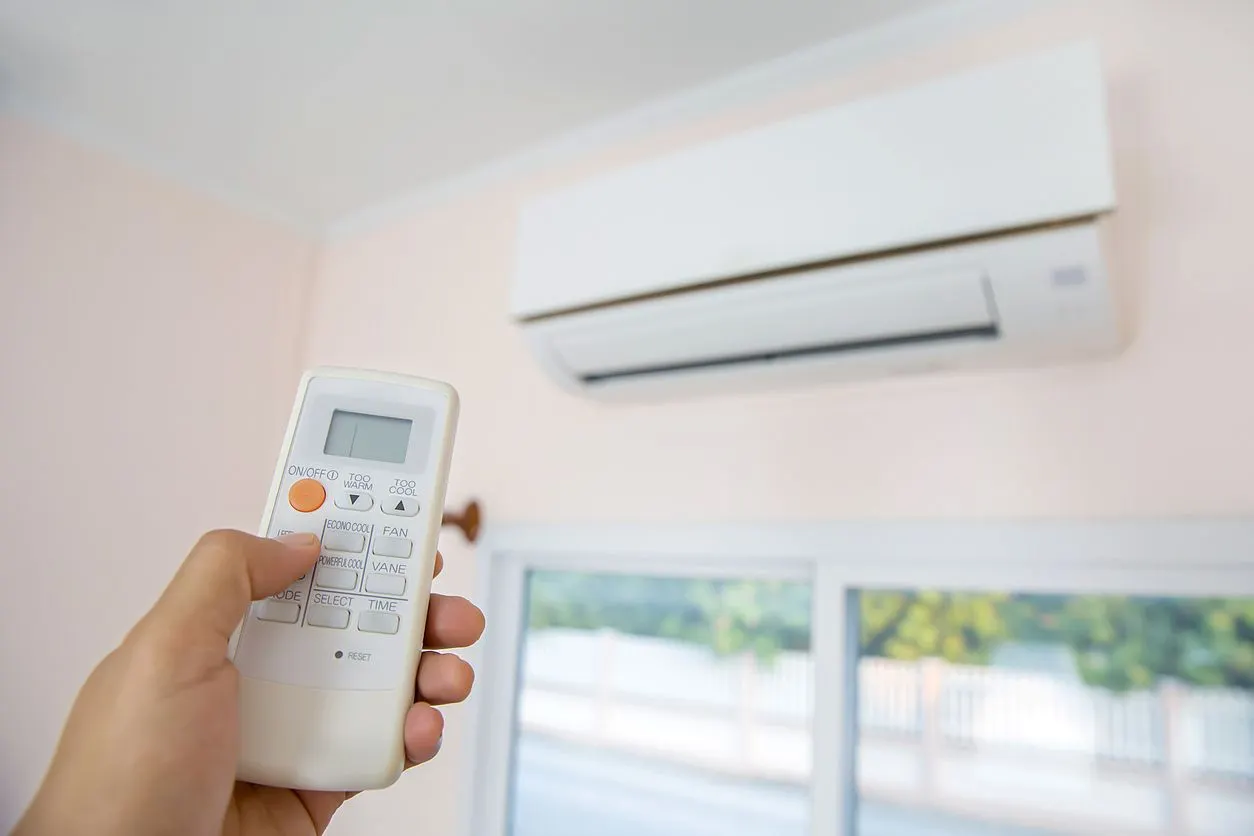 A hand holds a remote control aimed at a wall-mounted white air conditioner in a room.