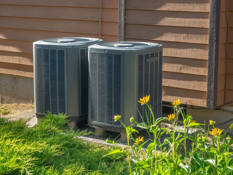 Two gray HVAC air conditioning units sit outdoors against a wooden house wall next to green foliage and yellow flowers.