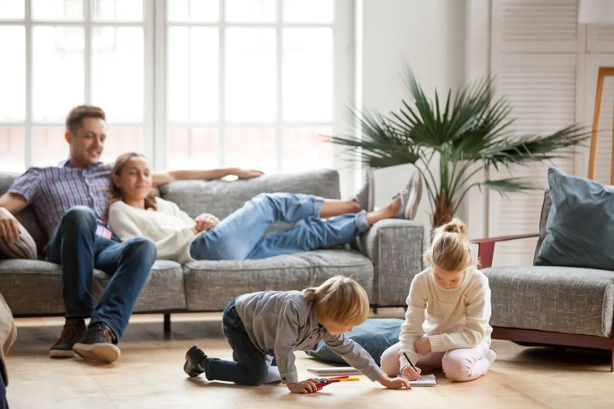 Parents relax on a sofa while two children play on the floor in a bright, modern living room.