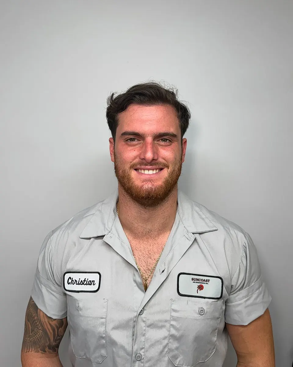 A person with a beard, wearing a short-sleeved light gray collared work shirt with patches, smiling against a gray wall.