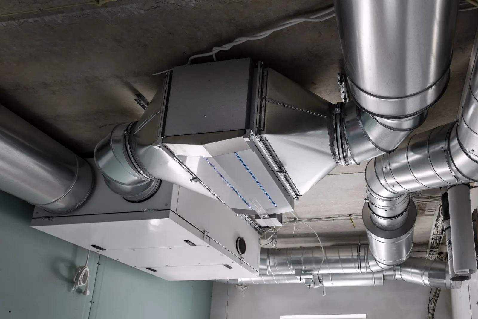 Metal ventilation ducts and a white HVAC unit mounted on a concrete ceiling.