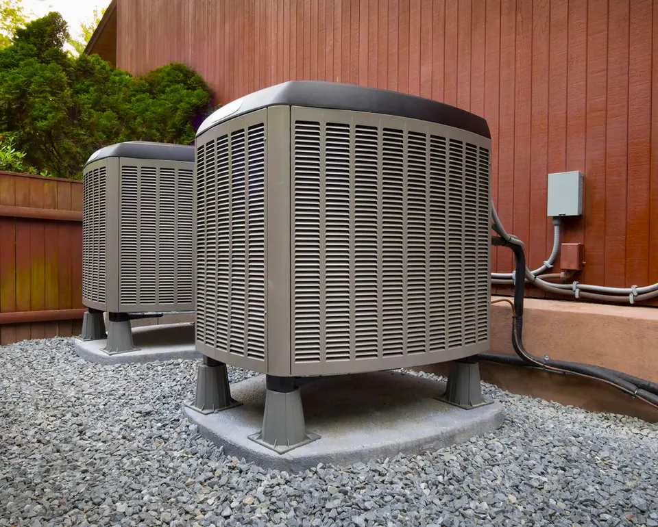 Two beige outdoor air conditioning units on concrete pads set on a gravel ground against a wooden wall.