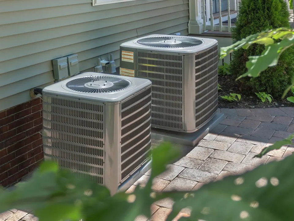 Two metallic residential air conditioning units sit on a paved surface next to a house with beige siding and brick.