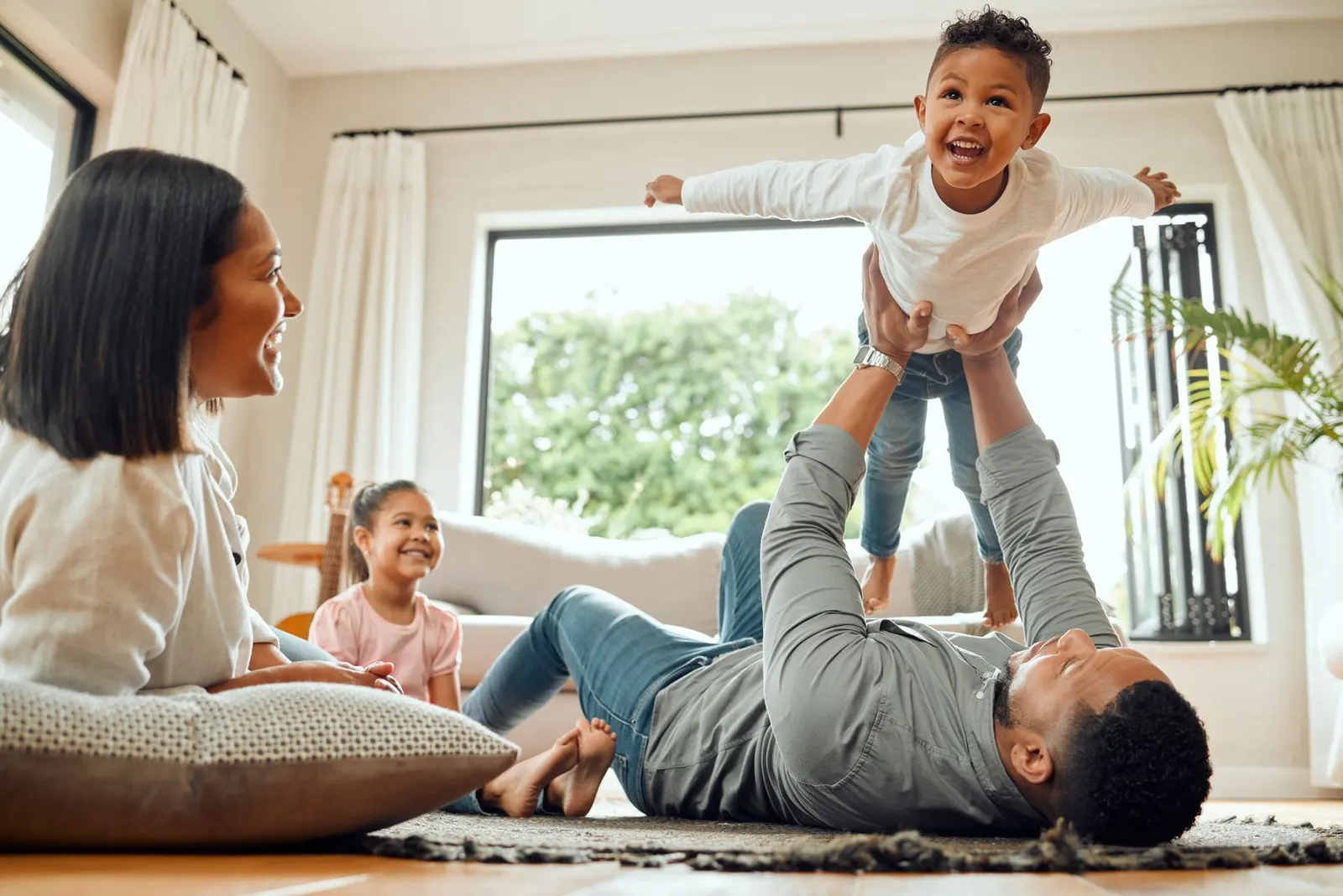 A parent lies on a living room rug, lifting a child into the air while another child watches and a person looks on.