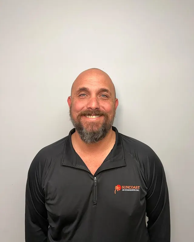 A smiling person with a beard, wearing a black long-sleeved athletic zip-up shirt against a plain light gray background.