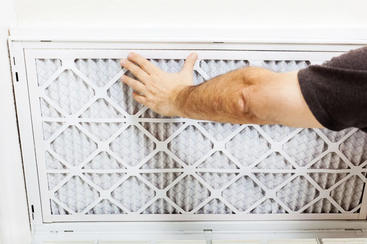 A person inserts a white pleated air filter into a residential wall-mounted return vent.