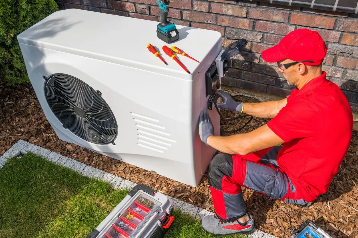A technician in a red uniform repairs an outdoor white heat pump unit with tools nearby in a yard.