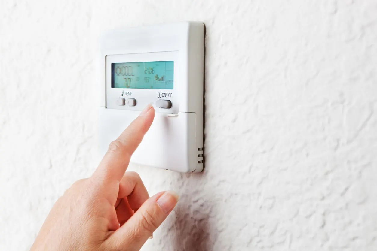 A finger presses a button on a wall-mounted digital thermostat.