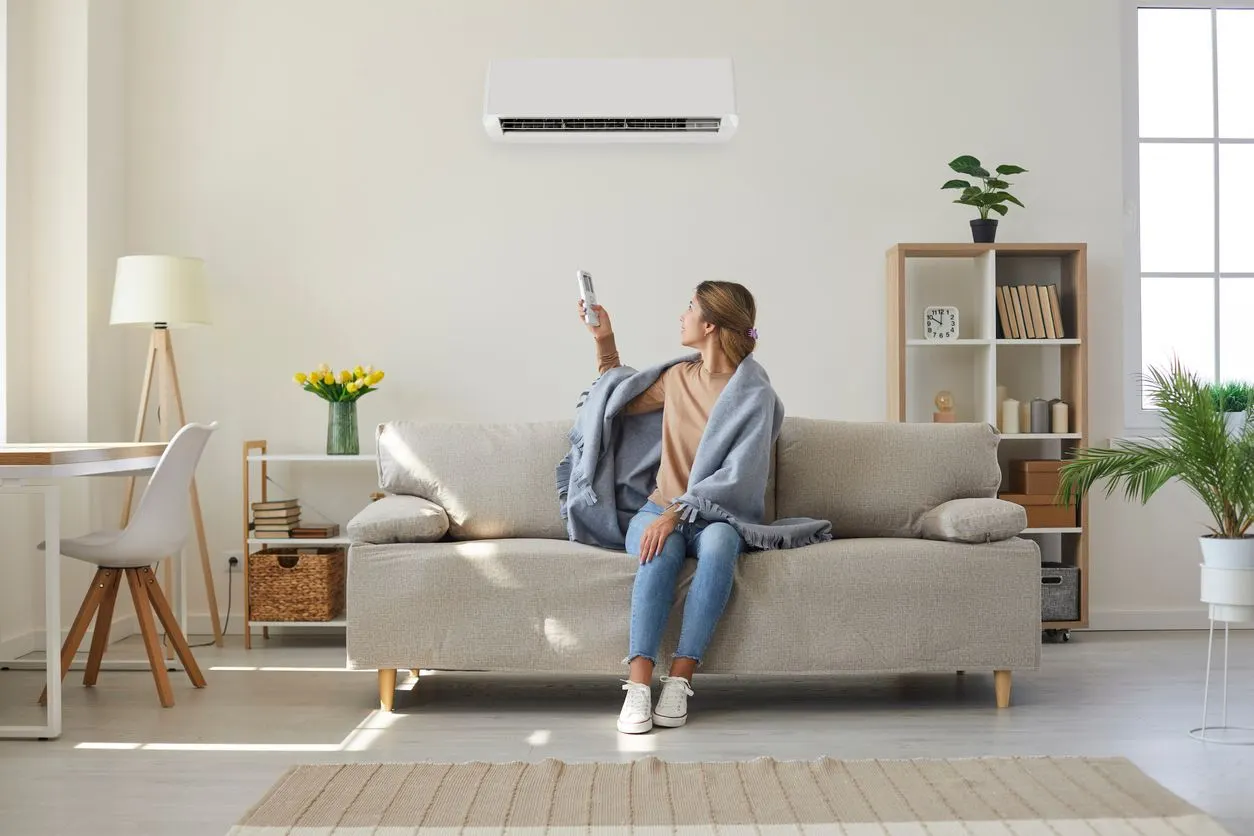 A person wearing a blanket sits on a sofa, pointing a remote at a wall-mounted air conditioner in a bright living room.