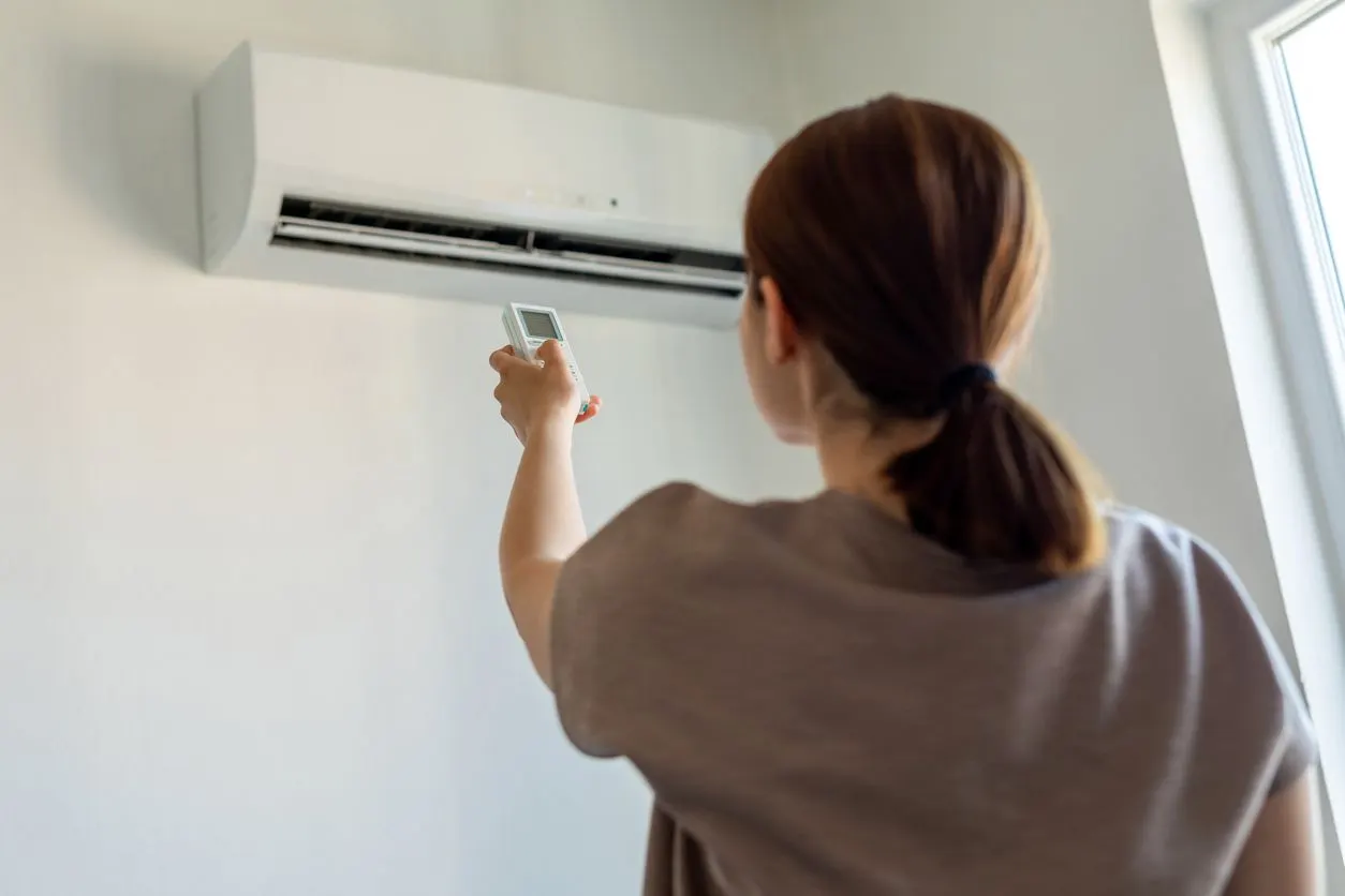 A person pointing a remote control at a wall-mounted air conditioning unit in a room.