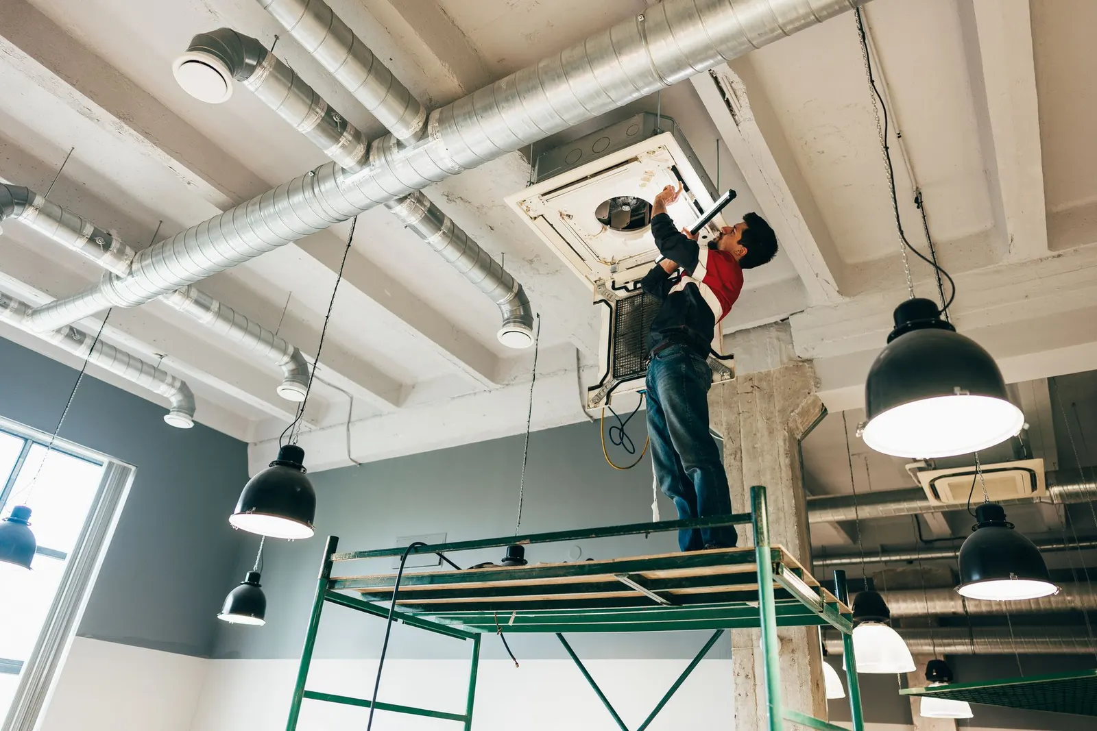 A person stands on scaffolding while repairing an air conditioning unit attached to an industrial ceiling with ductwork.